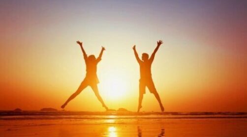 Two people in front of the sunset at the beach, jumping with their arms and legs out.