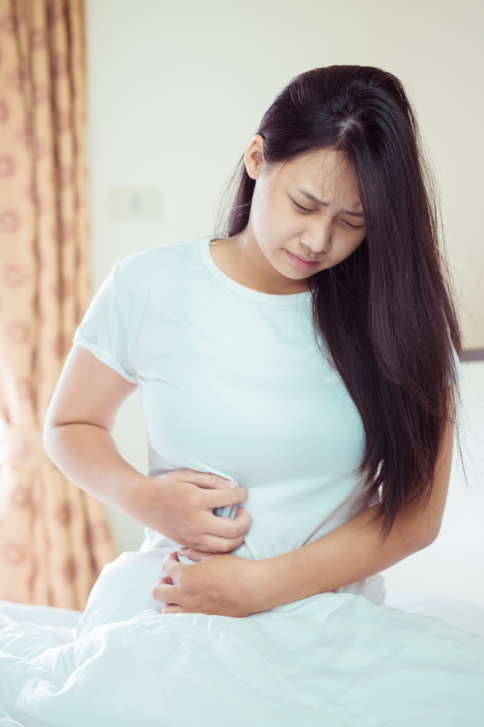 An Asian woman wearing white holding onto her stomach with her face showing pain.